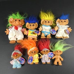 Puede incluir: Una colección de ocho muñecos Troll con varios colores de pelo, incluyendo verde, azul y amarillo. Los muñecos visten diferentes atuendos, como monos y vestidos. Cada muñeco tiene una etiqueta.