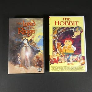 Peut inclure: Deux cassettes VHS sont présentées sur fond noir. La première est intitulée "The Lord of the Rings" avec une illustration fantastique. La seconde est intitulée "The Hobbit" avec une illustration de dessin animé d'un hobbit.