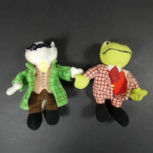 Puede incluir: Dos peluches: un tejón con un abrigo a cuadros verde y una rana con un chaleco rojo y una chaqueta a cuadros. El tejón tiene una máscara negra y la rana una corbata roja. Ambos tienen zapatos negros y se dan la mano.