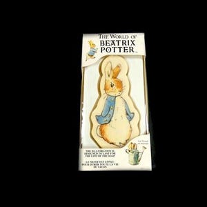 Puede incluir: Un jabón empaquetado con una ilustración de Peter Rabbit de The World of Beatrix Potter. El jabón tiene un fondo beige con un conejo de dibujos animados con una chaqueta azul. El texto en el empaque dice: "La ilustración está diseñada para durar toda la vida del jabón."