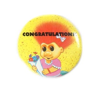 Könnte beinhalten: Ein runder gelber Ansteckbutton mit einem Cartoon-Troll mit leuchtend orangefarbenen Haaren, der ein rosa Kleid mit weißer Spitzenborte trägt. Der Troll hält einen Strauß bunter Blumen. Das Wort CONGRATULATIONS ist schwarz über dem Troll gedruckt.