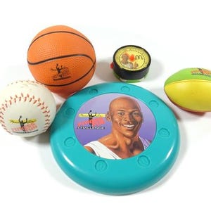 May include: A set of sports-themed toys including a basketball, baseball, a flying disc with a basketball player graphic, and a small round toy with a basketball player graphic. The flying disc is turquoise blue with the words "Dunkin' Challenge" printed on it.