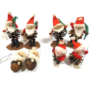 May include: A set of eight vintage Christmas ornaments featuring Santa Claus figures made from pine cones. The ornaments are dressed in red and white Santa suits with white beards and hats. Some of the ornaments are holding pine branches and others are holding skis.