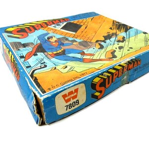 May include: A vintage Superman board game box with a blue background and a picture of Superman flying. The box has the text "Superman" and "Whitman" on it. The box is in good condition with some wear and tear.