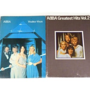 May include: Two vinyl record albums by ABBA. The album on the left is titled "Voulez-Vous" and features a blue and white geometric design with the band members standing in front of it. The album on the right is titled "ABBA Greatest Hits Vol. 2" and features a brown cover with a photo of the band members.