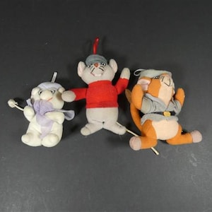 Può includere: Tre peluche di animali su una superficie scura. I giocattoli includono un topo bianco con una camicia rossa, un topo bianco con una sciarpa viola e un gatto arancione con un abito grigio. Ogni giocattolo ha un filo attaccato.
