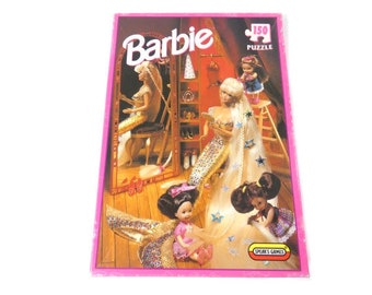 May include: A 150 piece Barbie jigsaw puzzle featuring three Barbie dolls. The dolls are wearing gold and white dresses with long hair. The puzzle box has a pink background with the words "Barbie" and "150 Puzzle" in white.