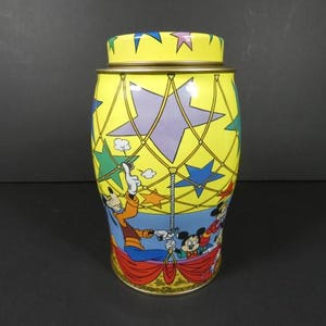 May include: A yellow, blue, and purple tin container with a design of Mickey Mouse and friends in a hot air balloon. The tin has a gold lid and is decorated with stars.