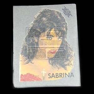 May include: A print featuring a woman with long, dark hair and striking eyes. The image has the name "SABRINA" at the bottom, with a cross and text overlay. The overall aesthetic is vintage, with a retro feel.