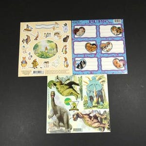 Op de afbeelding: Drie stickervellen op een zwart oppervlak. Eén vel toont cartoon dieren en een landschap. Een ander heeft hartvormige kaders met hondenafbeeldingen en het woord "PUPS". Het derde vel toont dinosaurus illustraties.