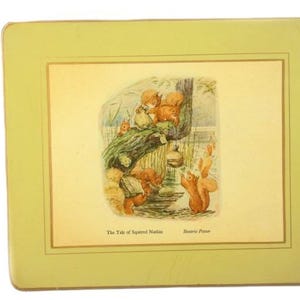 May include: A rectangular placemat with a light green border and a framed illustration. The illustration depicts squirrels in a forest setting, with text that reads "The Tale of Squirrel Nutkin" and "Beatrix Potter".