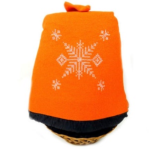 Puede incluir: Manta de punto naranja con un diseño de copo de nieve blanco. La manta está doblada y colocada en una cesta tejida.