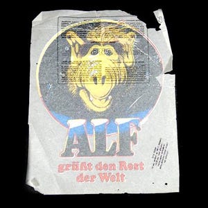 May include: Vintage paper featuring an ALF graphic. The image shows a yellow-faced alien character with the word "ALF" in blue and grey, and the German phrase "grüßt den Rest der Welt".