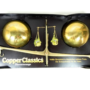 May include: A brass scale with two round bowls, a black base, and the words "Copper Classics Blumenwaage" printed on the front. The scale is a decorative item for the home.