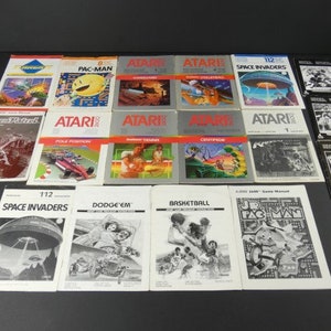 Puede incluir: Una colección de manuales de instrucciones de videojuegos Atari vintage, incluyendo Pac-Man, Space Invaders y Dodge 'Em. Los manuales son de varios colores y presentan ilustraciones de los juegos.