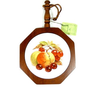 Puede incluir: Una tabla de queso octogonal de madera con un inserto de cerámica que presenta un diseño pintado de fruta, incluyendo melocotones, cerezas y hojas. La tabla tiene un mango de madera con un lazo para colgar.