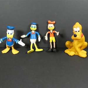 Könnte beinhalten: Vier Disney-Charakterfiguren: Donald Duck, ein weiterer Donald Duck, eine dritte Figur und Pluto. Donald Duck trägt ein blaues Matrosenhemd und eine blaue Mütze. Der andere Donald Duck trägt ein blaues Hemd und eine Fliege. Die dritte Figur trägt ein orangefarbenes Hemd und gelbe Shorts. Pluto ist gelb.