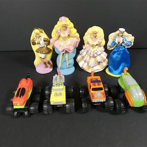 Puede incluir: Una colección de figuras de Barbie vintage y camiones monstruo de juguete. Las figuras de Barbie están en varios vestidos y poses, con cabello rubio. Los camiones de juguete son rojos, amarillos, naranjas y verdes, con ruedas negras.