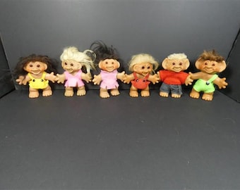 May include: A set of six troll dolls with distinct hair colors and clothing. The dolls feature tan skin, pointed ears, and wide smiles. They are dressed in a yellow outfit, a pink dress, a pink jumper, a red outfit, a red sweater with jeans, and green overalls.