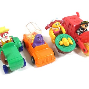 Op de afbeelding: Een set van vier speelgoedvoertuigen met personages uit populaire kinderprogramma's. De voertuigen zijn een groene tractor met een clown achter het stuur, een oranje jeep met een paarse monster achter het stuur, een rode vrachtwagen met een gestreept personage achter het stuur en een geel en rood voertuig met een personage dat een mand met gele aardappelen vasthoudt.