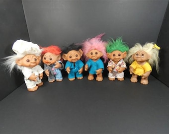 May include: A collection of six troll dolls with distinct hair colors including white, red, black, pink, green, and blonde. Each doll features a unique outfit, such as a chef hat and uniform, blue overalls, a blue shirt, a patterned shirt, and a yellow top.