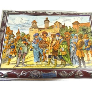 May include: A vintage tin box with a colourful illustration of people in medieval clothing standing in front of a castle. The text on the box reads "Meister Erwin erhielt von Nürnberg's Rathen und Rathsherrn einen neuen und erstandenen Geldbus."
