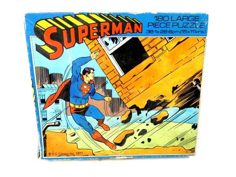 May include: A vintage Superman 180-piece puzzle box with a colorful illustration of Superman flying through the air. The box is blue with yellow and red stripes and features the text "SUPERMAN 180 LARGE PIECE PUZZLE 38-1x28-6cm.(15x11%ins.)" and "DC Comics Inc. 1977".