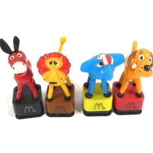 May include: Set of four plastic finger puppets featuring a red donkey, a yellow lion, a blue elephant, and an orange dog. Each puppet is on a square base with the letter "M" printed on it.