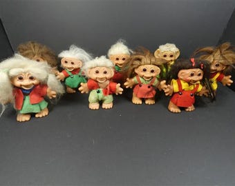 May include: A collection of eight small troll dolls with various hair colors including white, brown, and black. Each doll features a distinct facial expression, pointed ears, and is dressed in colorful fabric outfits such as red, green, and yellow.
