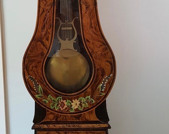 May include: A tall, antique, wooden grandfather clock with a painted floral design. The clock face has Roman numerals and a gold-colored frame. The clock has a pendulum and a glass door revealing the inner workings.