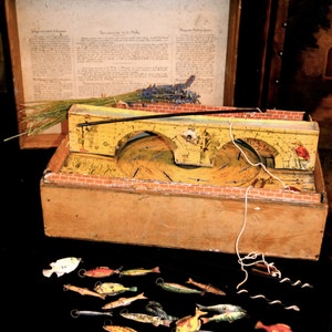 May include: An antique wooden game with a yellow bridge design, a wooden box, and a collection of small, colorful fish. The game includes a printed document and a sprig of lavender. The game is likely a vintage fishing game.