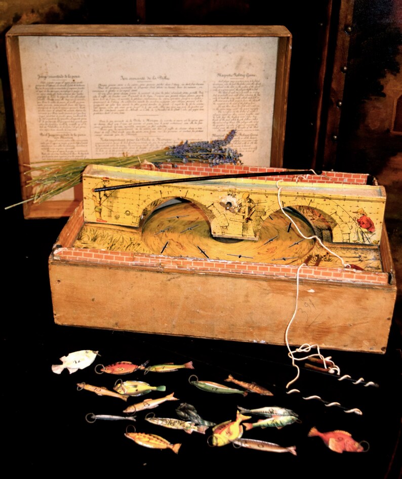 May include: An antique wooden game with a yellow bridge design, a wooden box, and a collection of small, colorful fish. The game includes a printed document and a sprig of lavender. The game is likely a vintage fishing game.