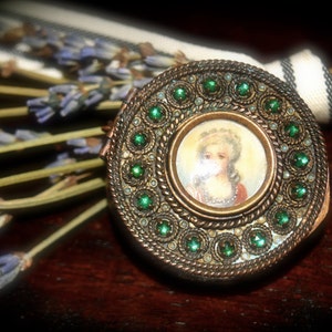 Puede incluir: Un medallón redondo de color dorado con un borde de piedras preciosas verdes. El medallón tiene un pequeño retrato ovalado de una mujer con un cuello de encaje blanco y un vestido azul claro.