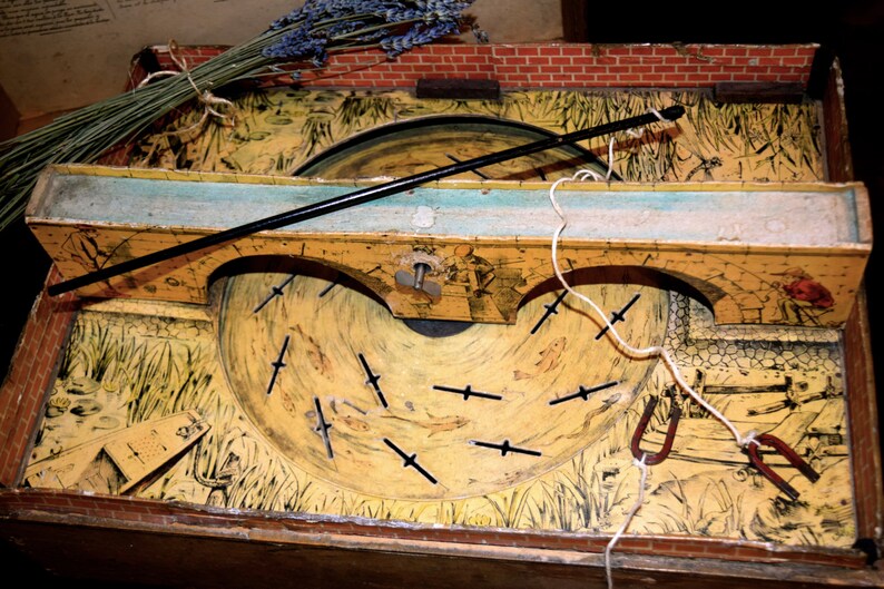 May include: Vintage wooden fishing game with a painted landscape scene. The game features a rotating disc with fish cutouts, a black fishing rod, and a brick border. A bundle of dried lavender rests on top.
