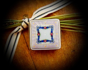 Könnte beinhalten: Eine kleine, quadratische, weiße Emaille-Box mit einem Blumendesign in Blau, Rosa und Gold. Die Box hat einen dekorativen Rand und steht auf einem Holzgrund.