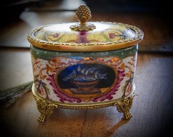 May include: A decorative porcelain trinket box with a gold trim and a painted scene of birds in a bowl. The box has a lid with a gold finial and sits on three gold feet.