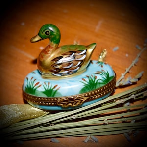 Könnte beinhalten: Eine Porzellan-Schmuckdose in Form einer Ente. Die Ente ist mit grünen, goldenen und weißen Details bemalt. Die Dose ist mit grün und gold gemalten Schilf und einem goldenen Rand verziert.