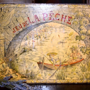 May include: Vintage rectangular tin box with a painted illustration of a fishing scene. The words "JEU DE LA PECHE" are arched across the top. The artwork depicts a stone bridge, boats, and figures fishing in a river.