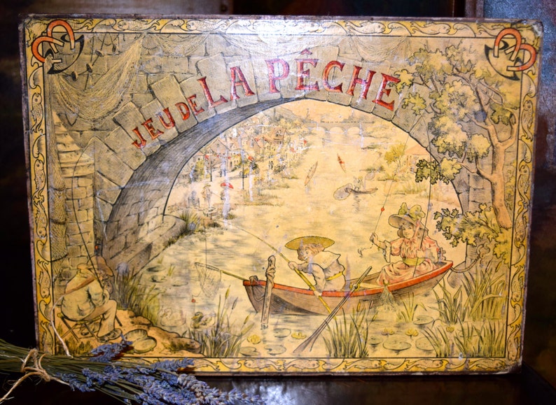 May include: Vintage rectangular tin box with a painted illustration of a fishing scene. The words "JEU DE LA PECHE" are arched across the top. The artwork depicts a stone bridge, boats, and figures fishing in a river.