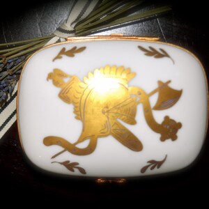 May include: A white, rectangular trinket box with a gold rim and a gold emblem of a helmet and axe. The box is decorated with gold leaf accents. Dried lavender and a striped ribbon are in the background.