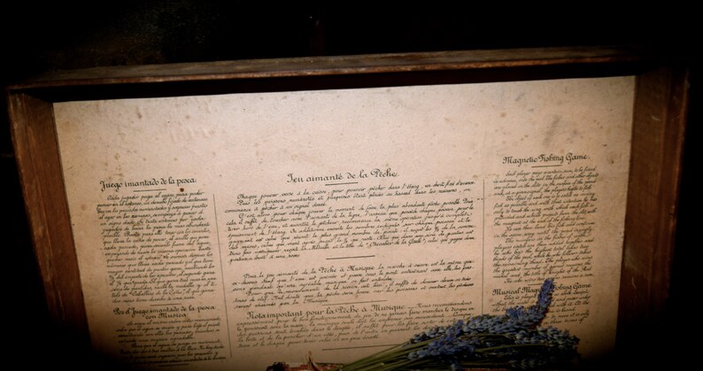 May include: A vintage wooden box displays a faded document with handwritten text in multiple languages. The document is aged, with visible wear and tear. A sprig of lavender rests at the bottom, adding a touch of color and texture to the scene.