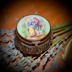 May include: A small, round, antique trinket box with a hinged lid. The lid is decorated with a painted scene of two people in a garden setting. The box is made of brass and has an ornate design.