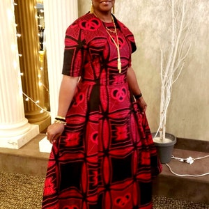 May include: A woman wearing a red and black patterned maxi dress with a gold necklace and earrings. She is standing in front of a white tree and a gold column.