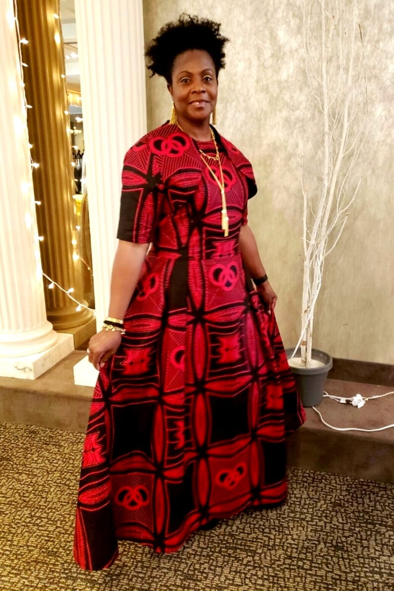 May include: A woman wearing a red and black patterned maxi dress with a gold necklace and earrings. She is standing in front of a white tree and a gold column.