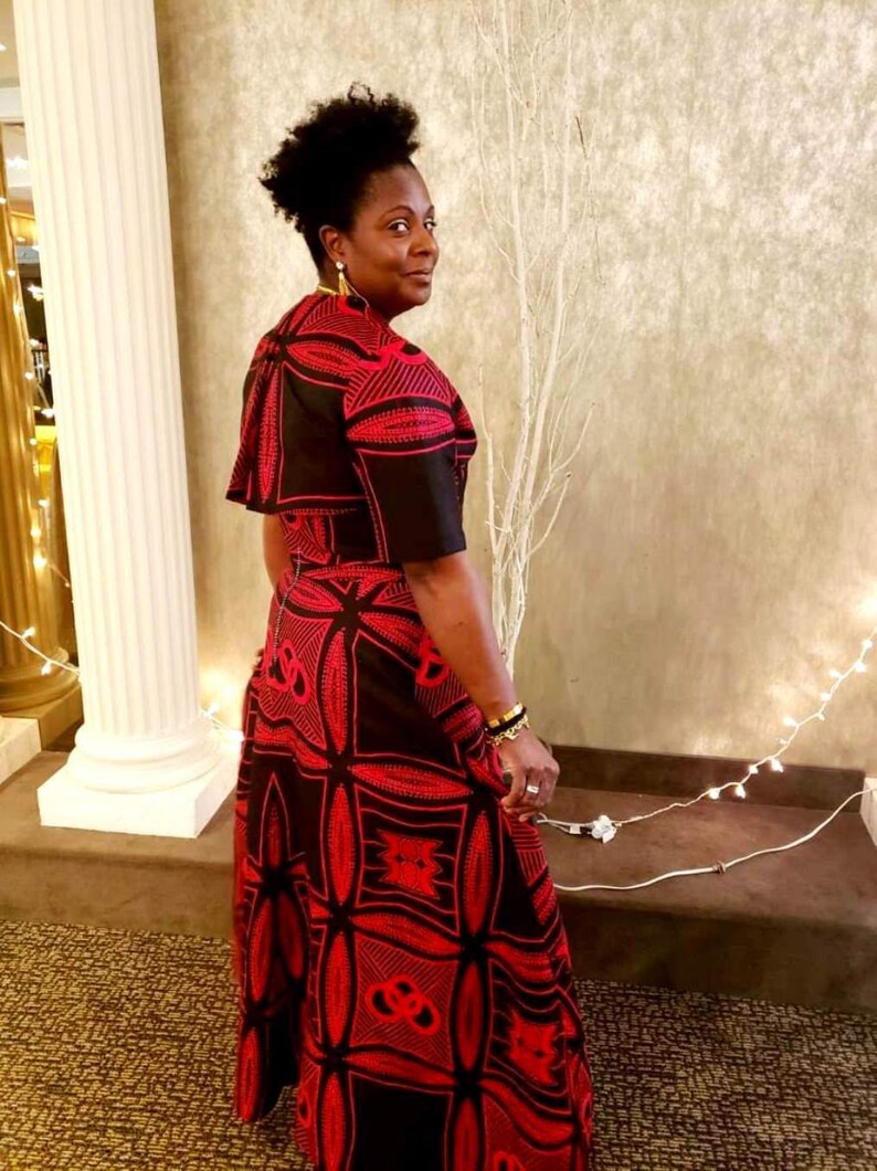 May include: A woman wearing a black and red dress with a geometric pattern. The dress has a short sleeve and a long skirt. The woman is standing in front of a white wall with a white branch in the background.
