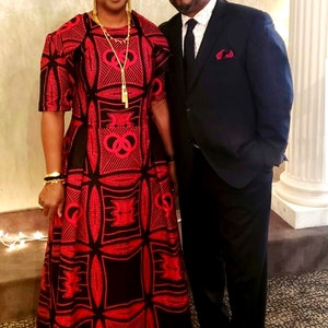 May include: A couple posing for a photo. The woman is wearing a red and black patterned dress with a gold necklace. The man is wearing a black suit with a white shirt and a black tie.
