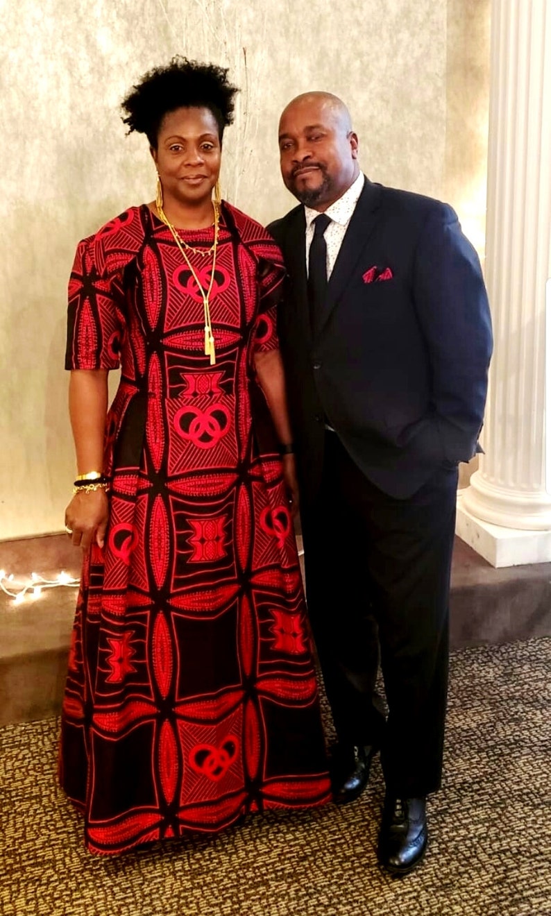 May include: A couple posing for a photo. The woman is wearing a red and black patterned dress with a gold necklace. The man is wearing a black suit with a white shirt and a black tie.