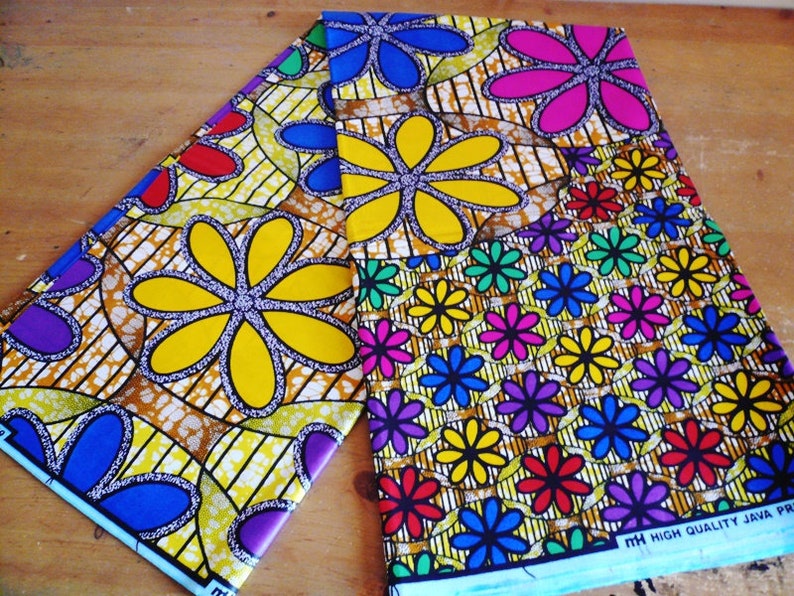 May include: Two pieces of colorful fabric with a floral pattern. The fabric has a yellow background with blue, red, green, purple, and pink flowers. The fabric is printed with the text "HIGH QUALITY JAVA PRINT".