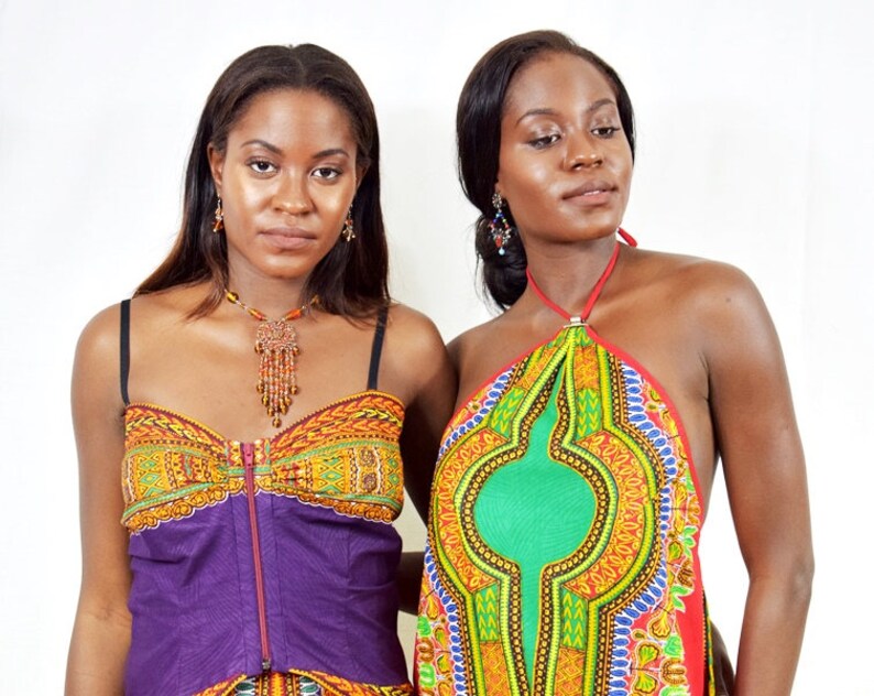 May include: Two women wearing colourful African print tops. One woman is wearing a purple top with a zipper and a beaded necklace. The other woman is wearing a halter top with a green, yellow and red pattern.