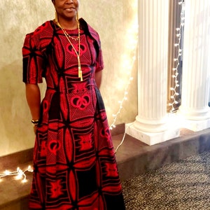 May include: A woman wearing a red and black patterned dress with a gold necklace. The dress has a fitted bodice and a full skirt. The woman is standing in front of a white wall with string lights.